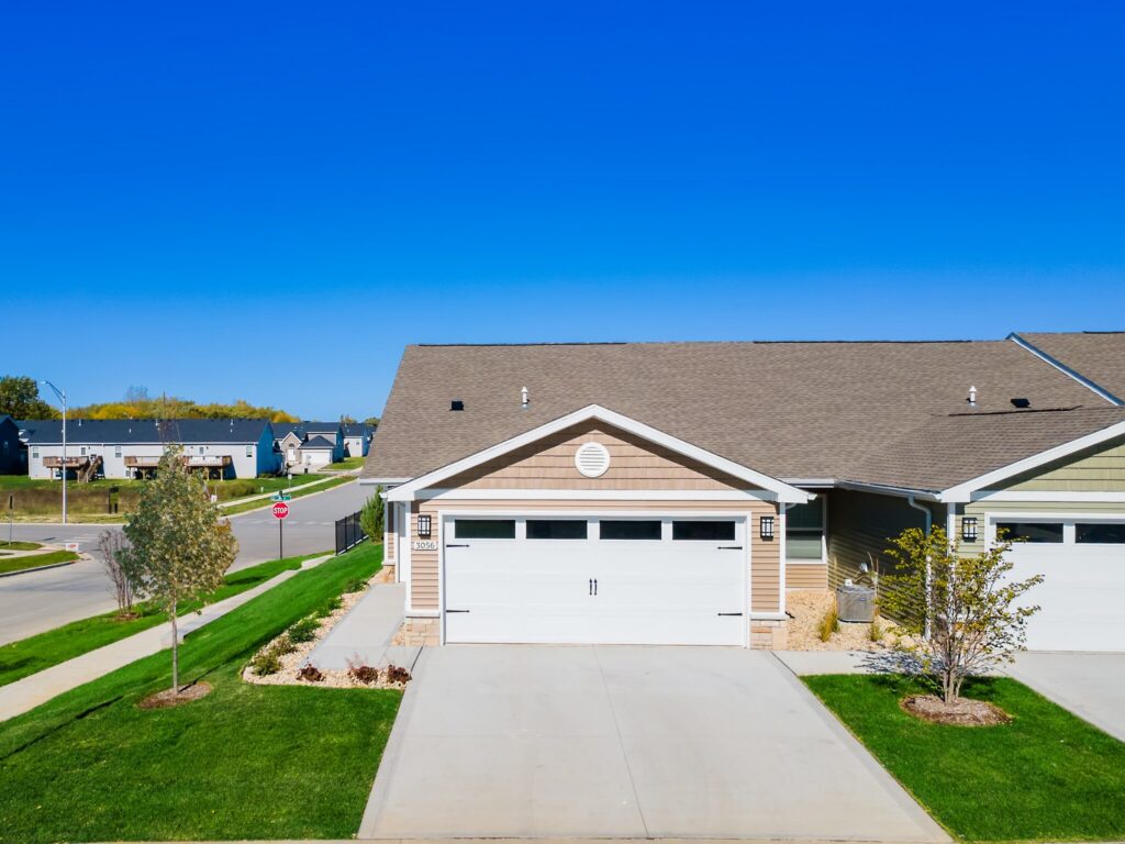Suburban duplex with two-car garage, beige siding, and a well-maintained lawn on a corner lot under a clear blue sky.