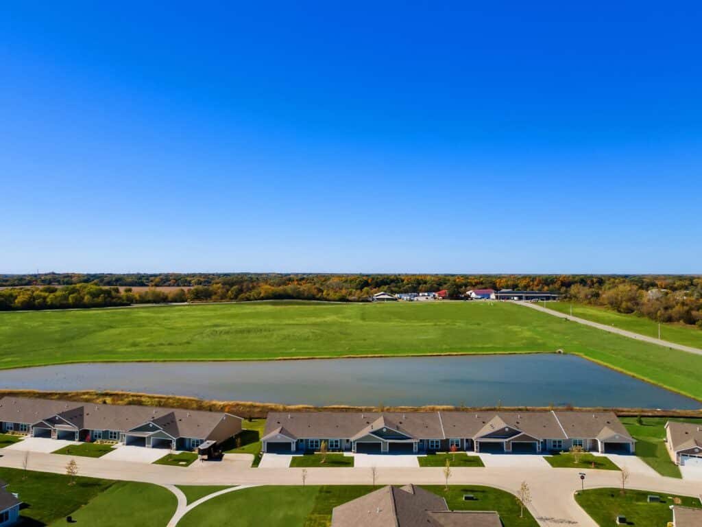 A row of single-story houses with driveways faces a pond and open green field under a clear blue sky.