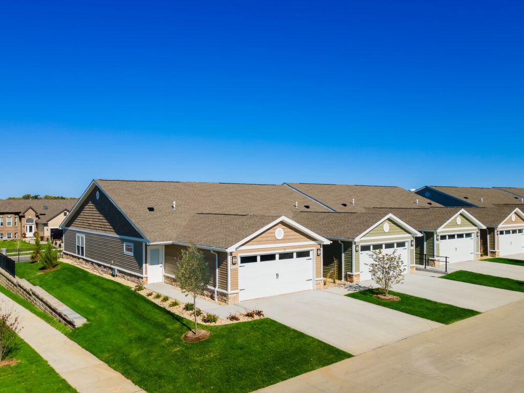 Row of single-story suburban houses with attached garages, green lawns, and a clear blue sky above.