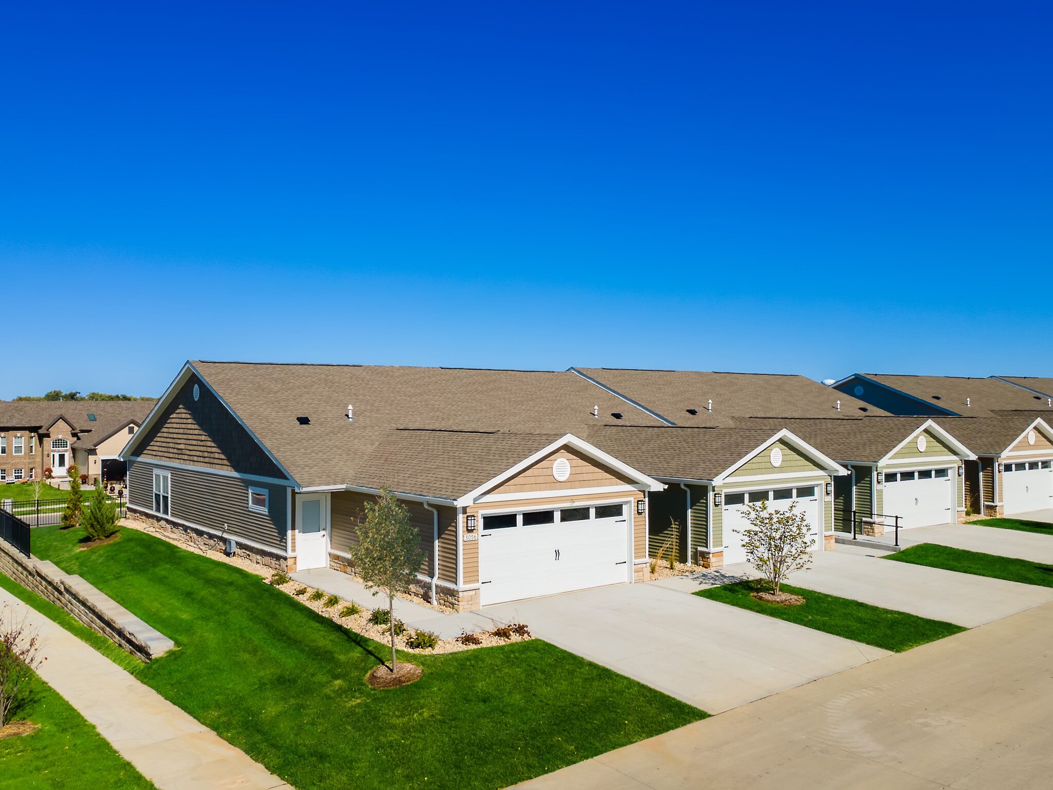 Row of single-story suburban houses with attached garages, green lawns, and a clear blue sky above.