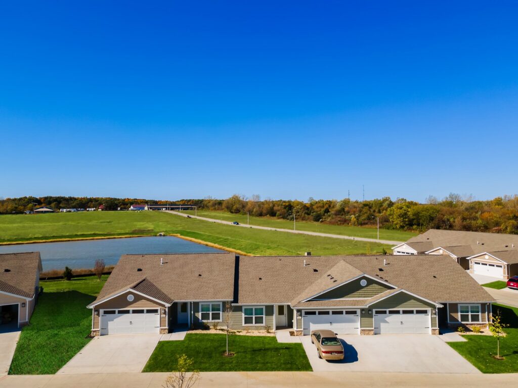Row of single-story houses with garages facing a driveway, a pond and green fields in the background under a clear blue sky.