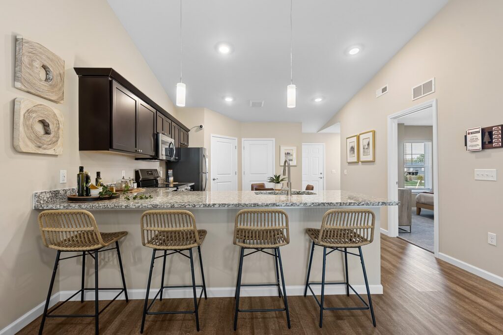Modern kitchen with a granite island, four wicker barstools, pendant lights, dark cabinets, and stainless steel appliances in an open-concept layout.