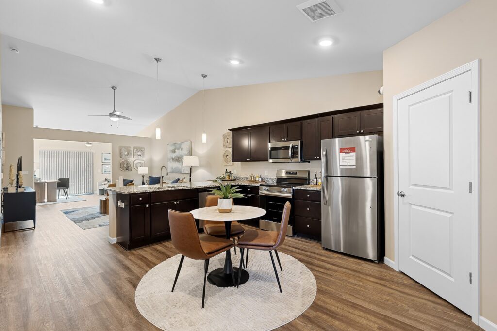 Modern kitchen and dining area with dark cabinets, stainless steel appliances, round dining table with two chairs, and wood flooring in an open-concept apartment.