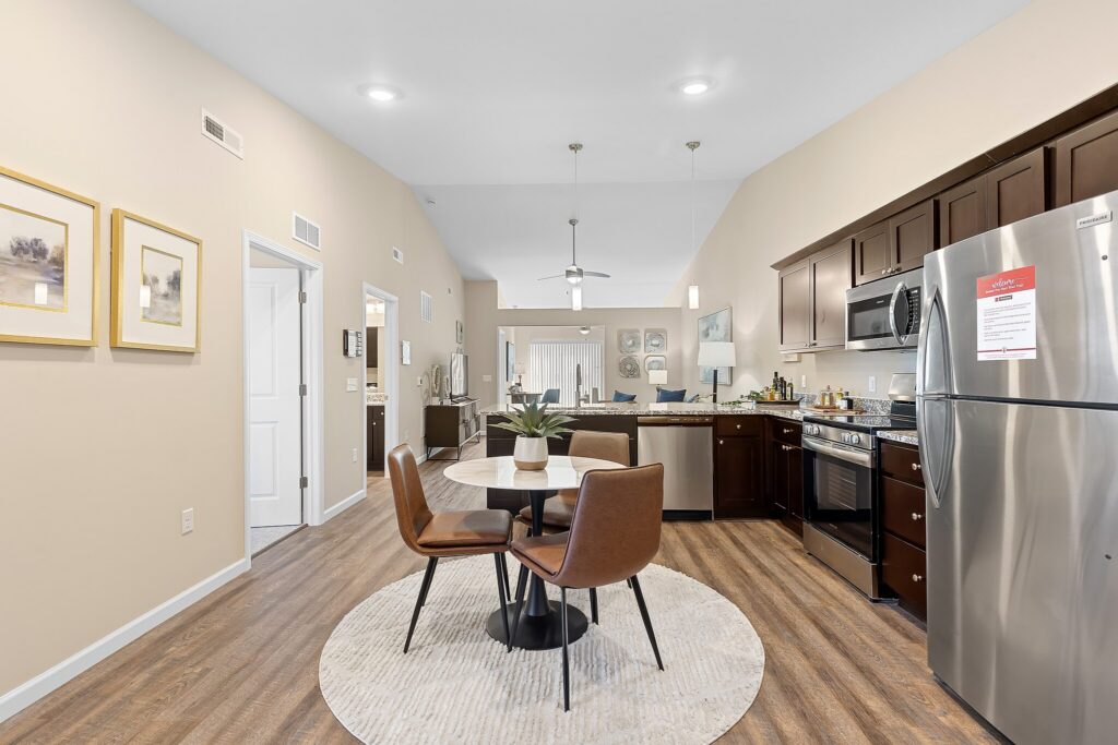 Modern kitchen and dining area with dark wood cabinets, stainless steel appliances, a round table with four brown chairs, and light-colored walls and flooring.