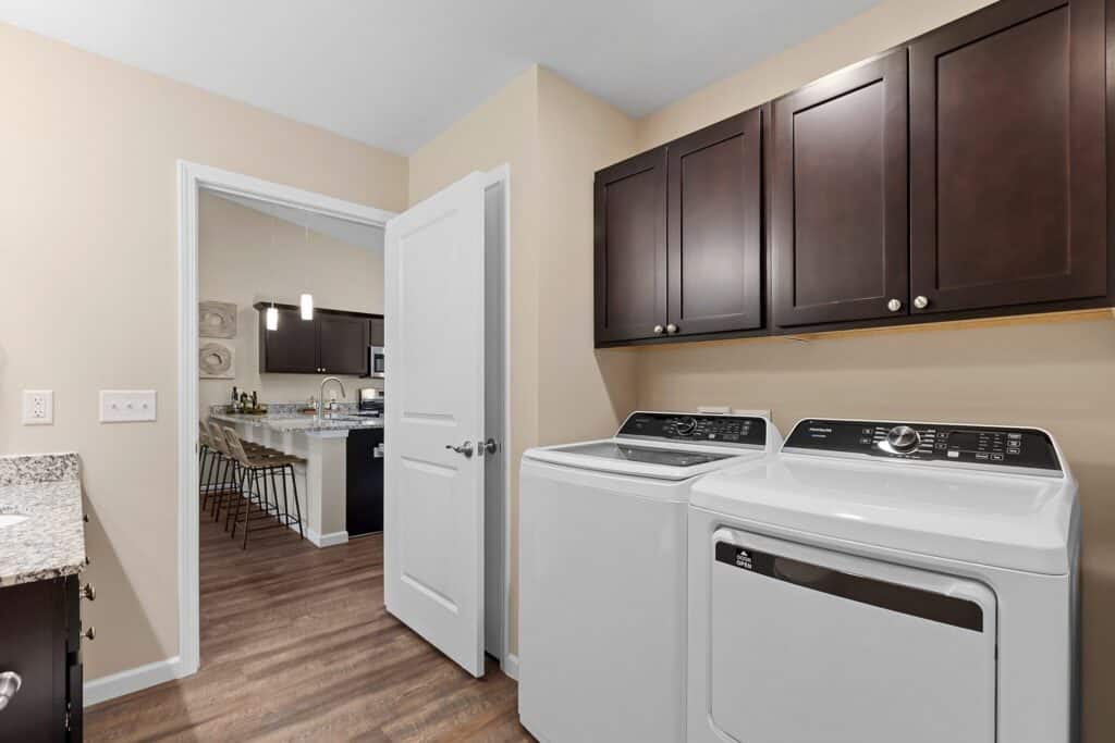 A laundry room with a washing machine and dryer, dark wood cabinets above, granite countertop, and an open door leading to a kitchen with bar stools.