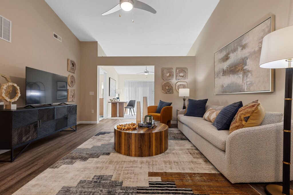 Modern living room with a beige sofa, abstract art, round wooden coffee table, TV on a black console, floor lamp, and a patterned rug. Dining area is visible in the background.