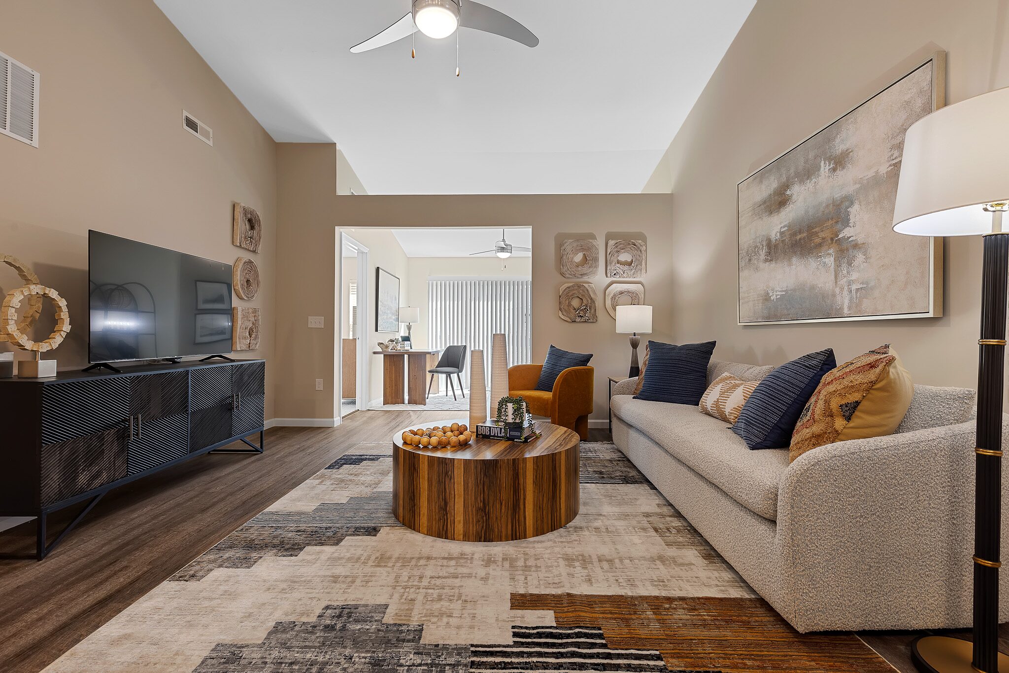 Modern living room with a beige sofa, abstract art, round wooden coffee table, TV on a black console, floor lamp, and a patterned rug. Dining area is visible in the background.