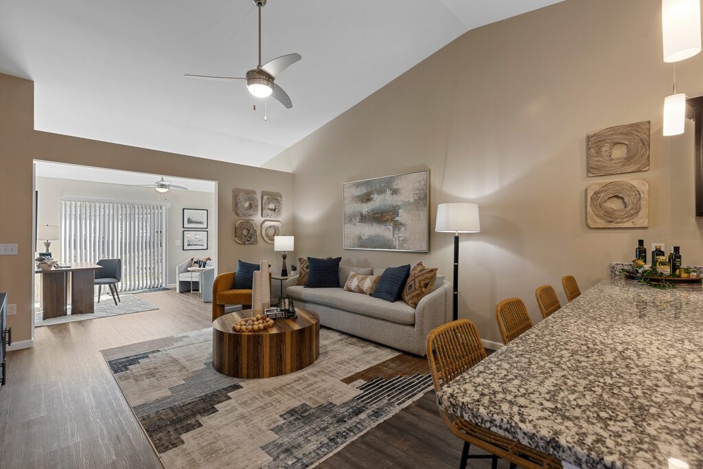 Modern living room with a beige sofa, wooden coffee table, area rug, and a granite kitchen island with barstools. A home office is visible in the background through an open doorway.