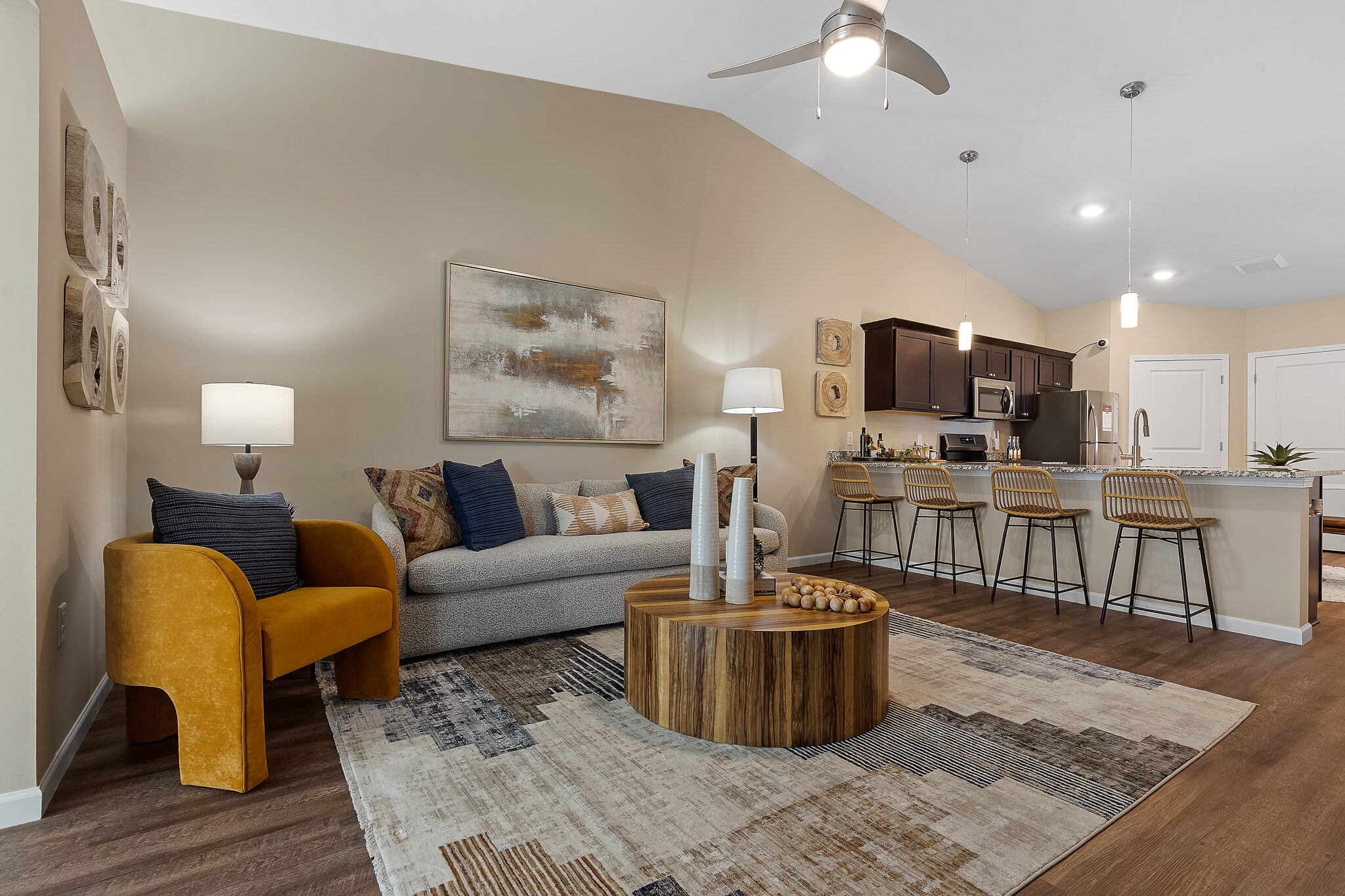 Modern living room and kitchen with neutral tones, a round wooden coffee table, bar stools at a granite island, and contemporary art on the walls.