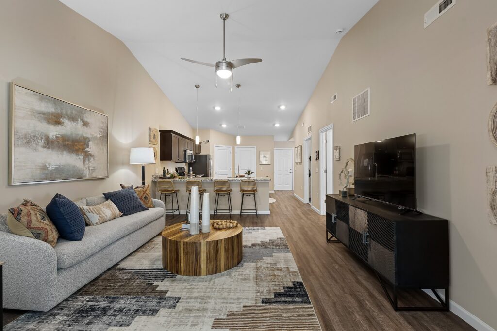 Modern open-concept living room and kitchen with neutral colors, a gray sofa, wooden coffee table, bar stools, TV, and contemporary decor.