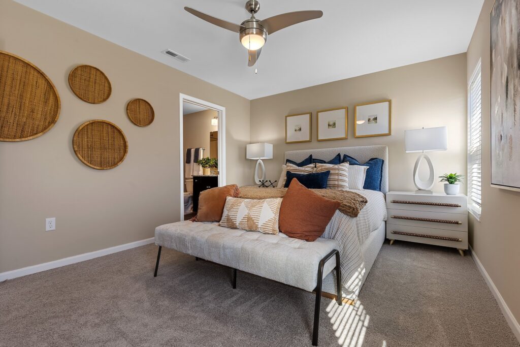 A modern bedroom with a neatly made bed, decorative pillows, a bench at the foot, two lamps on nightstands, and woven wall decor. Light beige walls and gray carpet flooring.