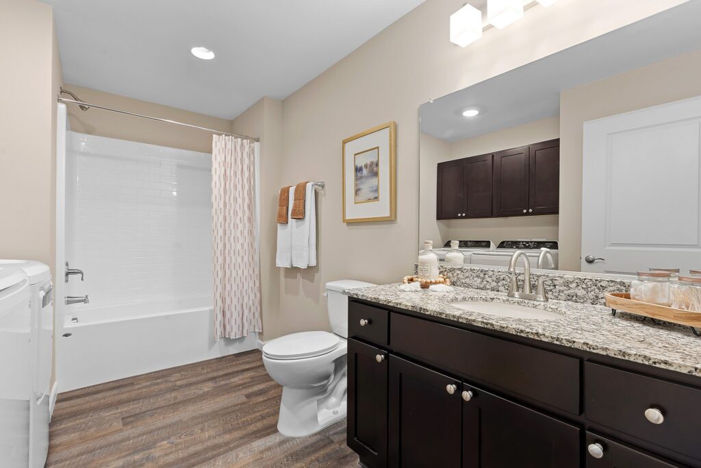 Modern bathroom with granite countertop, dark wood cabinets, a toilet, washer, bathtub with white tiles, and a shower curtain; towels and framed art on the wall.