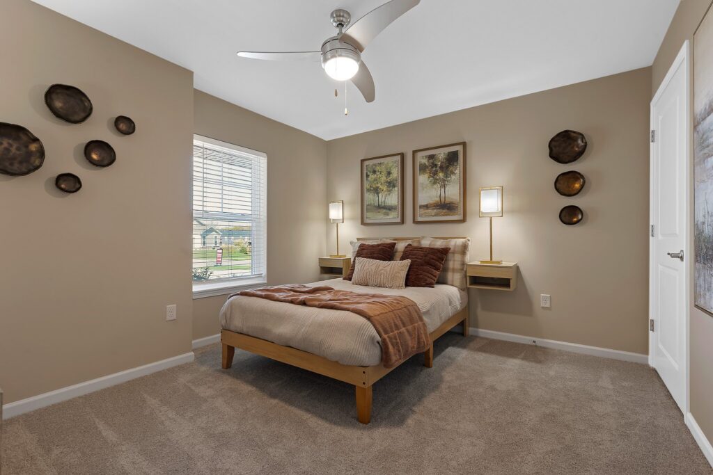 A neatly made bed with brown and beige bedding sits between two floating nightstands and lamps in a carpeted bedroom with tan walls and decorative wall art.