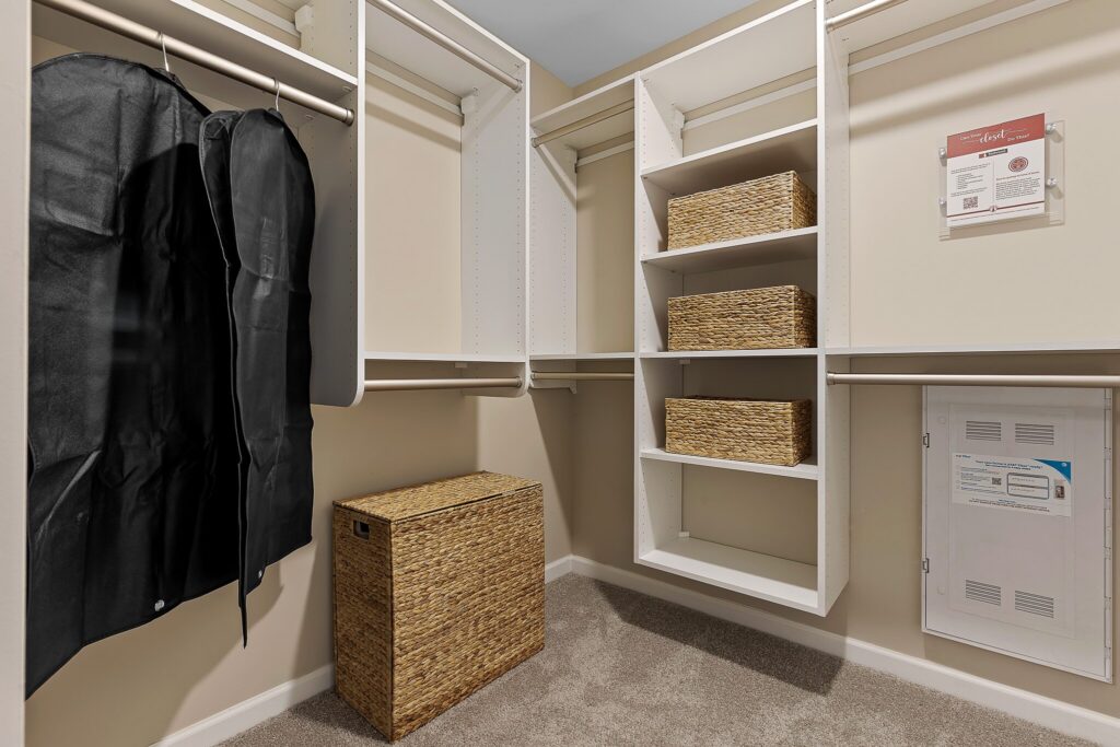 Walk-in closet with white shelves, hanging rods, four woven baskets, two black garment bags, a woven hamper, and utility panels on a beige carpet floor.