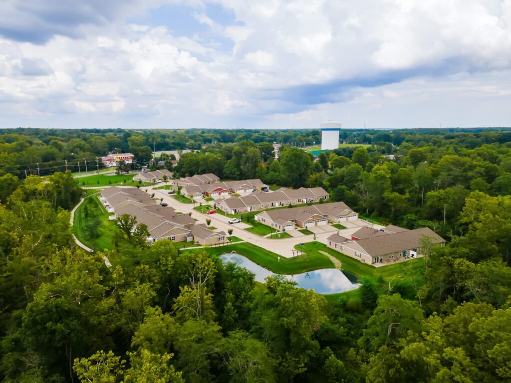 Aerial view of a residential housing complex surrounded by dense green trees, with a small pond and a large water tower in the background under a cloudy sky.