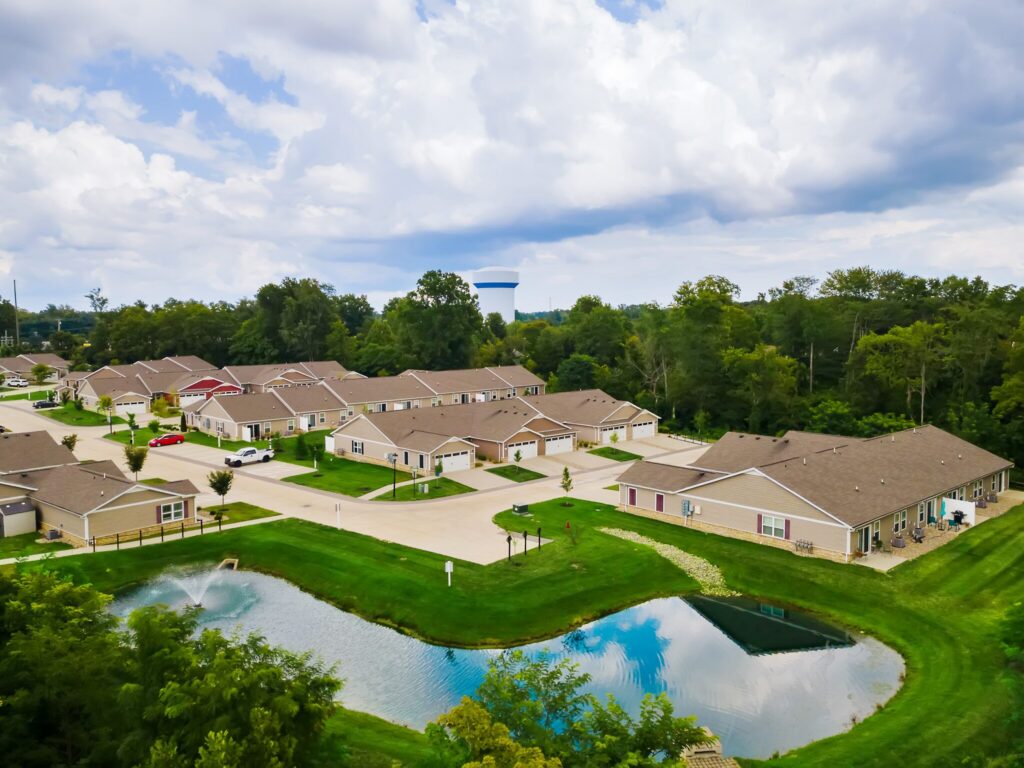 Aerial view of a suburban neighborhood with single-story houses, driveways, green lawns, a small pond, and a water tower in the background.