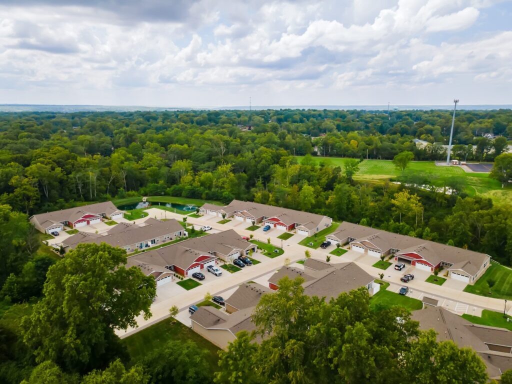 Aerial view of a suburban neighborhood with rows of single-story houses, surrounded by trees and greenery under a partly cloudy sky.