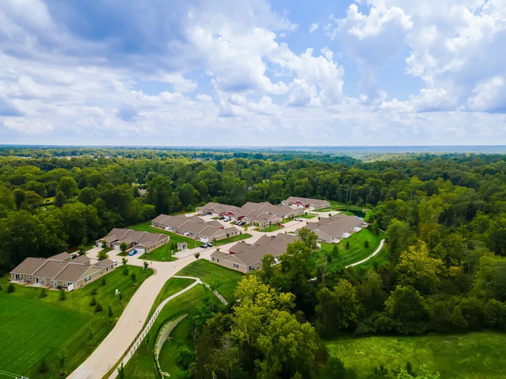 Aerial view of a suburban neighborhood with rows of houses surrounded by green trees and lawns under a partly cloudy sky.