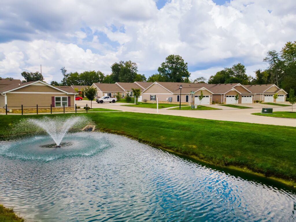 A small pond with a fountain sits in front of a row of single-story houses in a suburban neighborhood under a partly cloudy sky.