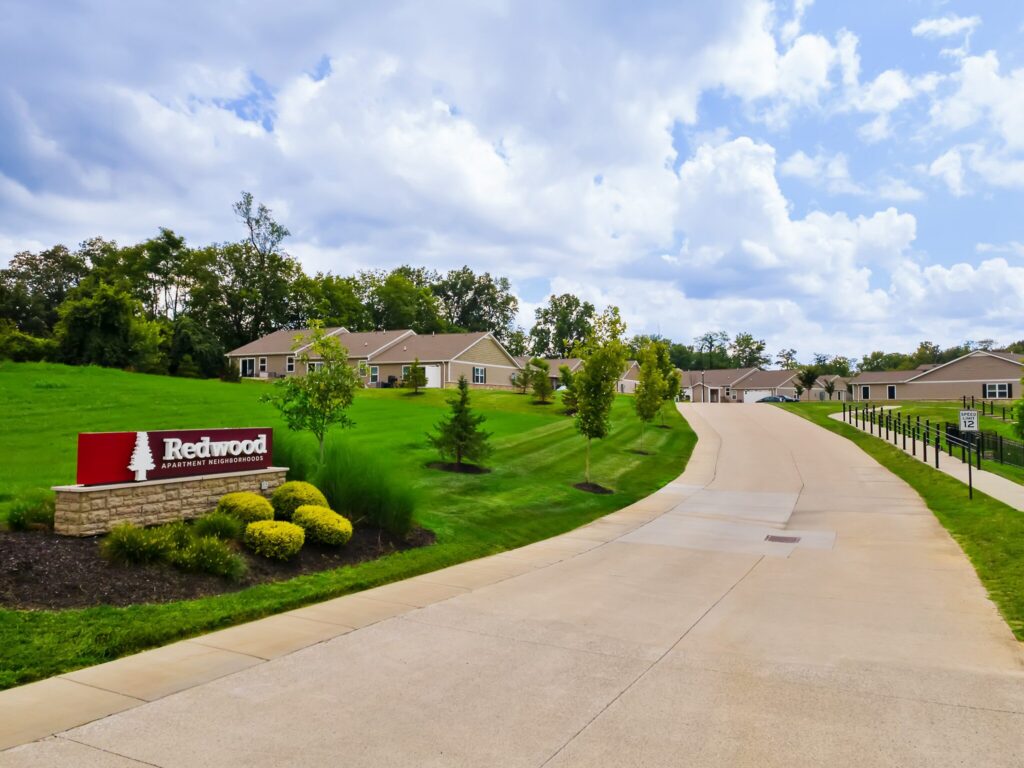 A paved driveway leads to single-story apartment buildings with a "Redwood Apartment Neighborhoods" sign on a landscaped lawn under a partly cloudy sky.