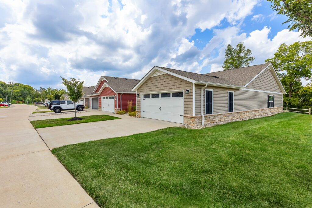 A row of modern single-story houses with garages and well-kept lawns under a partly cloudy sky in a suburban neighborhood.