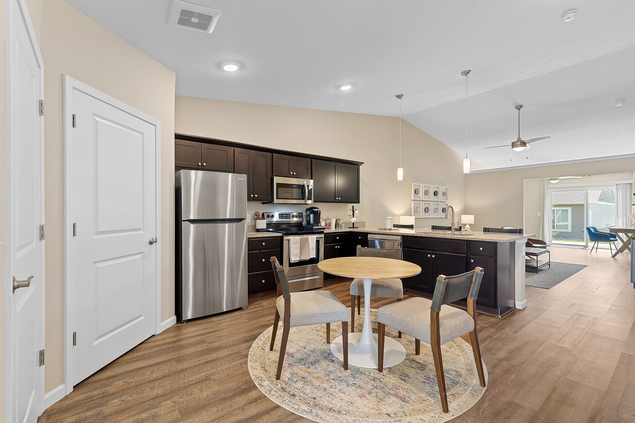 Modern kitchen with stainless steel appliances, dark cabinets, round dining table with four chairs on a rug, and open living area in the background.