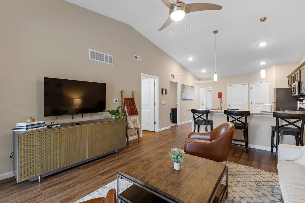 Modern living room and kitchen with beige walls, wooden floors, brown leather chairs, wall-mounted TV, and a breakfast bar with three stools under pendant lights.