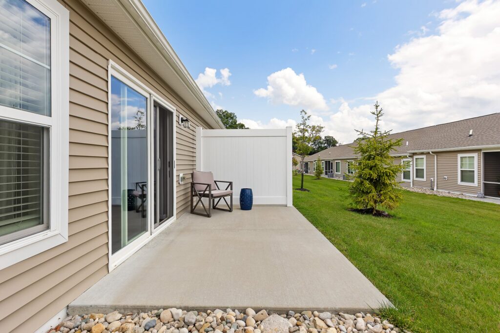 A concrete patio with a brown chair, small table, and blue planter sits outside a beige house with sliding glass doors, overlooking a grassy yard and neighboring houses.