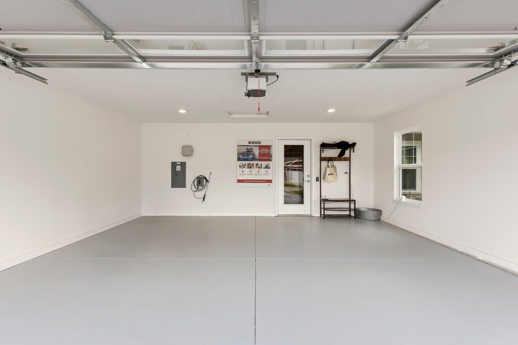 Clean, empty two-car garage with grey floors, white walls, ceiling lighting, wall-mounted utility panel, shelving unit, and a door with glass panes leading to another room.