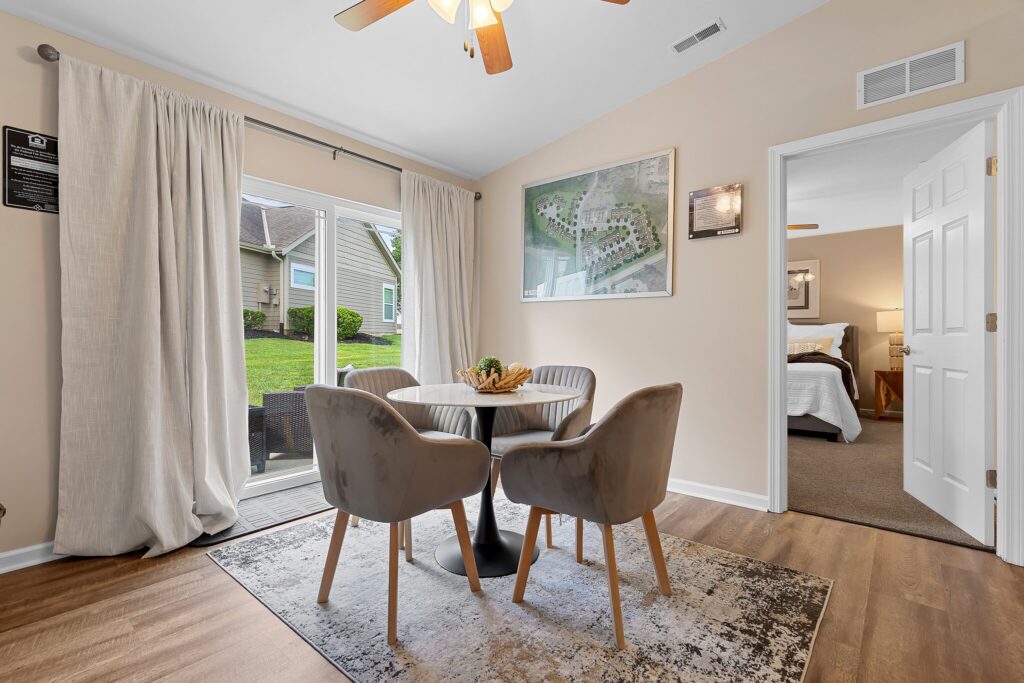Dining area with a round table and two gray chairs on a rug, next to glass doors leading outside; bedroom visible through an open door.