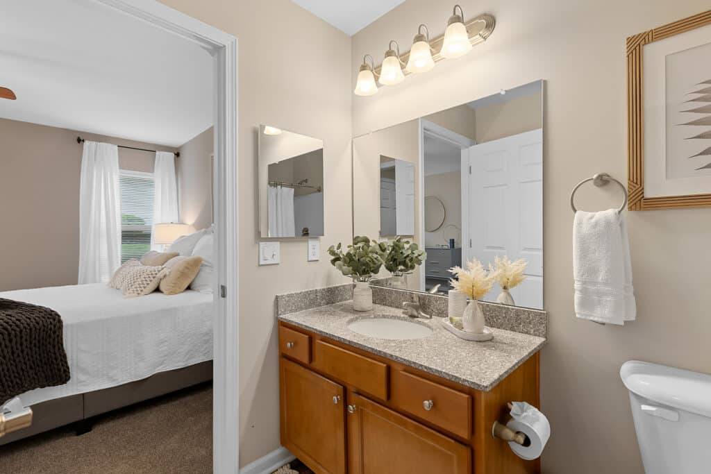 A bathroom with a granite countertop, wooden vanity, two mirrors, decorative plants, and a towel, adjacent to a bedroom with a made bed and window.