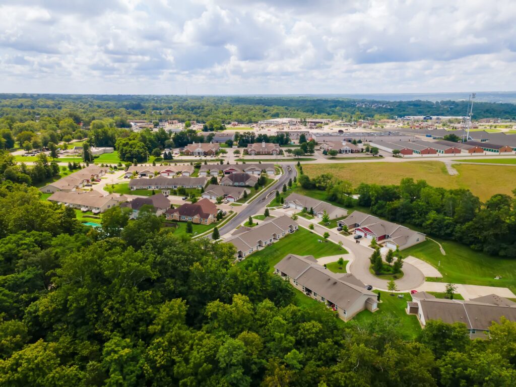 Aerial view of a suburban neighborhood with houses, roads, trees, and open fields under a partly cloudy sky.