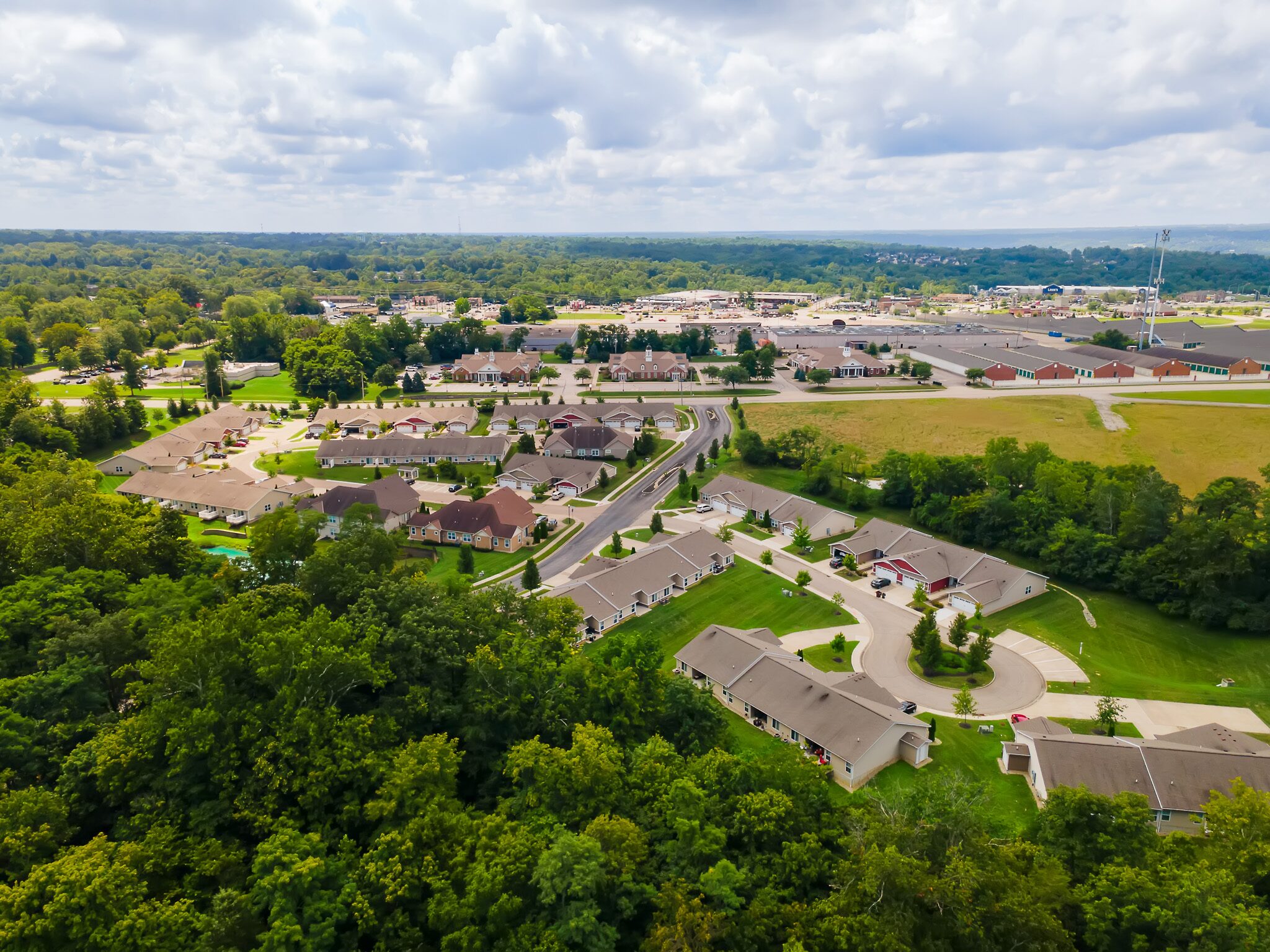 Aerial view of a suburban neighborhood with houses, roads, trees, and open fields under a partly cloudy sky.