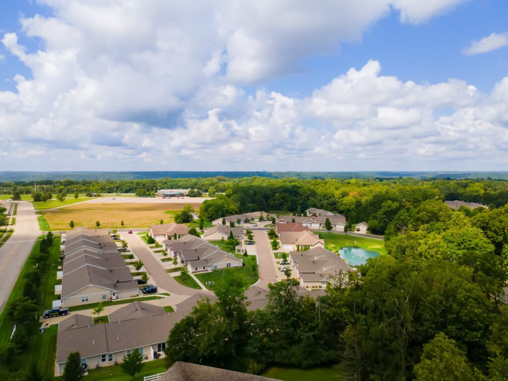 Aerial view of a suburban neighborhood with rows of houses, tree-lined streets, a small pond, and surrounding green landscape under a partly cloudy sky.