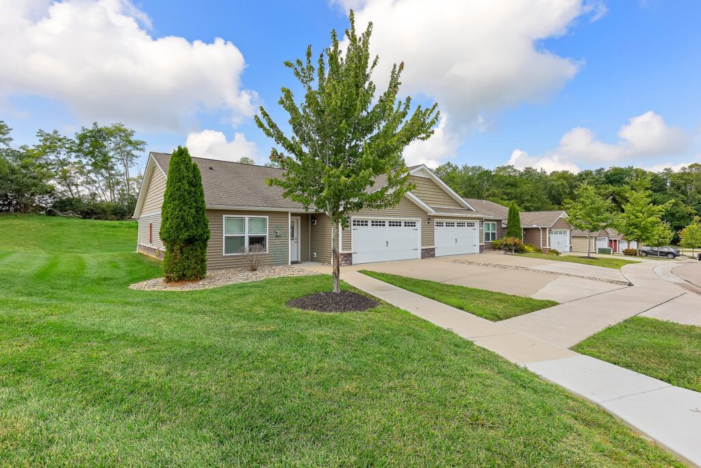 Single-story townhouse with attached two-car garage, manicured lawn, and a tree in the front yard, located on a corner lot in a suburban neighborhood.
