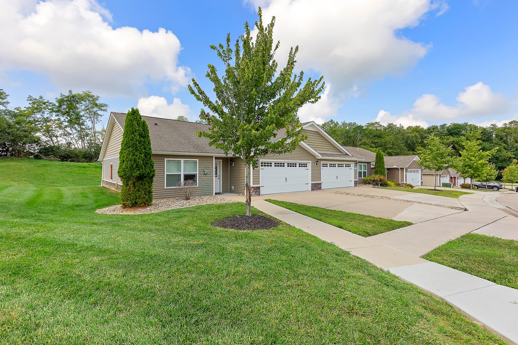 Single-story townhouse with attached two-car garage, manicured lawn, and a tree in the front yard, located on a corner lot in a suburban neighborhood.