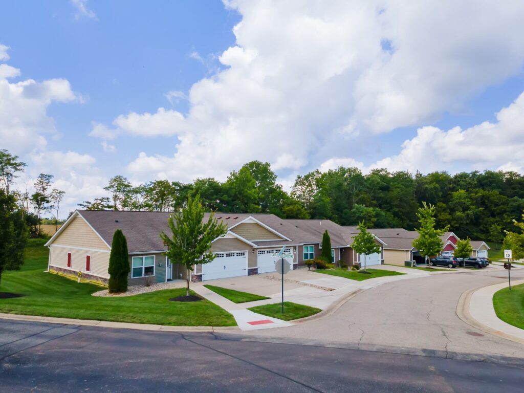 A row of single-story suburban houses with attached garages lines a curved residential street bordered by grass and trees under a partly cloudy sky.