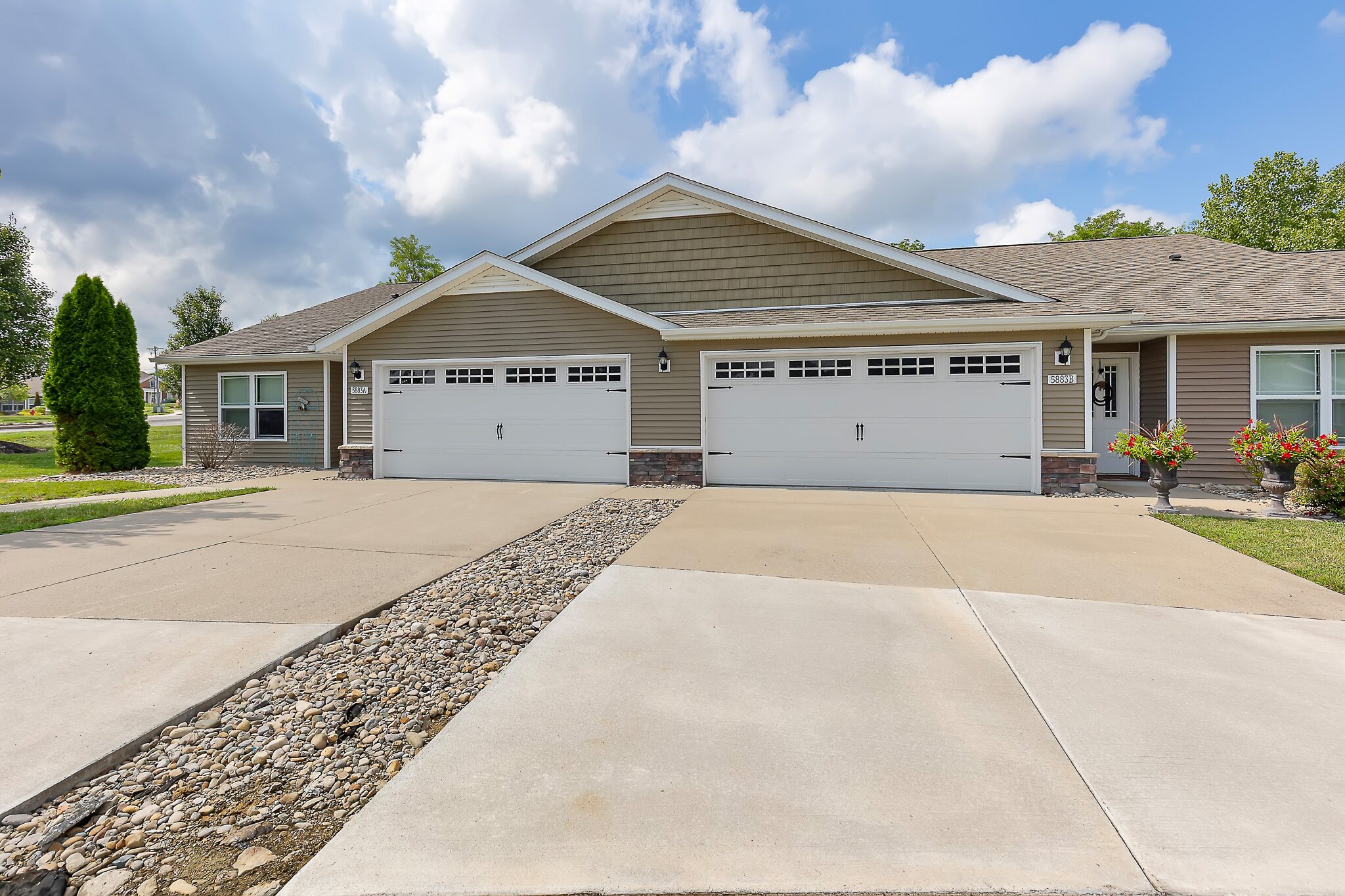 A single-story duplex with beige siding, stone accents, and two white double garages, separated by a rock-lined median on a concrete driveway.