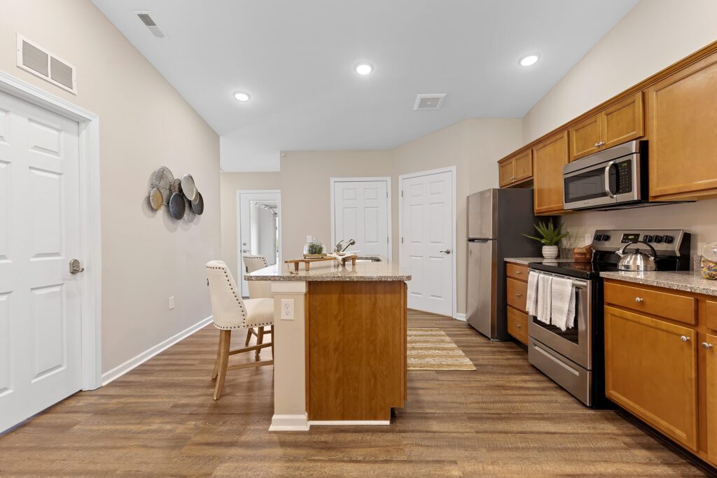 Modern kitchen with wood cabinets, stainless steel appliances, island with sink, two upholstered chairs, and decorative wall art, featuring hardwood flooring and neutral walls.