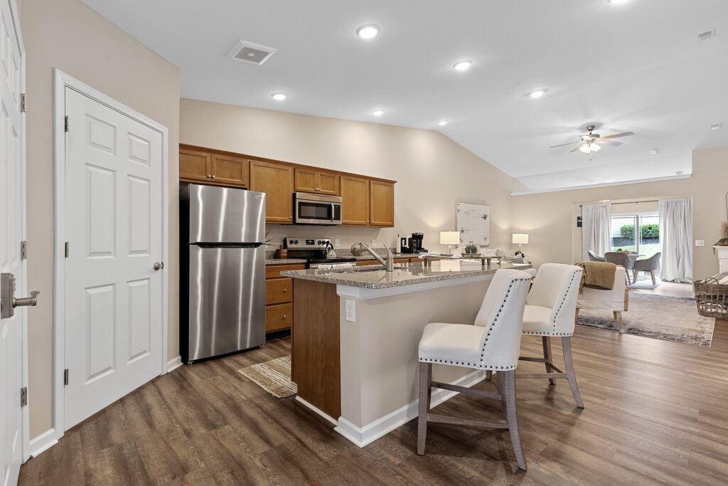 Modern kitchen with stainless steel appliances, wooden cabinets, island with two upholstered chairs, and open living area with sofa and sliding glass doors.