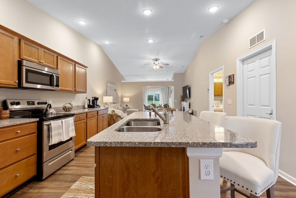 Modern kitchen with wooden cabinets, stainless steel appliances, granite island with double sink, and two cushioned stools; open layout leads to living area and patio doors.
