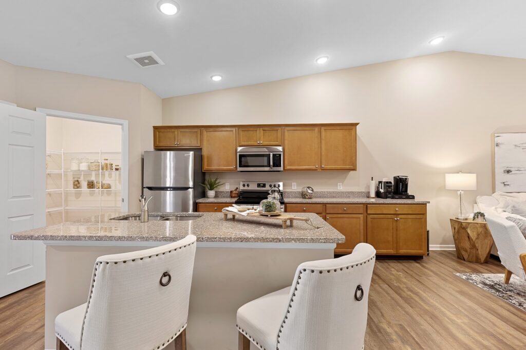 Modern kitchen with stainless steel appliances, wooden cabinets, a granite island with two white upholstered chairs, and a view into a pantry on the left.