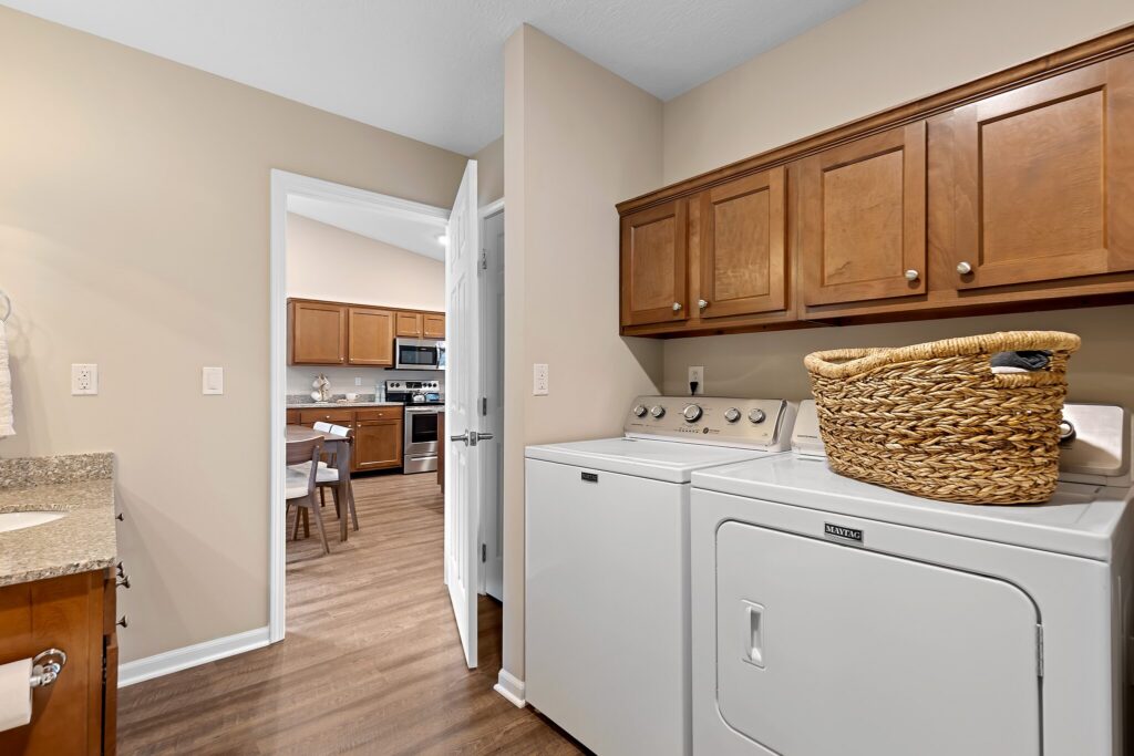 Laundry room with washer and dryer, wooden cabinets above, woven basket on dryer, and view into a kitchen with stainless steel appliances.