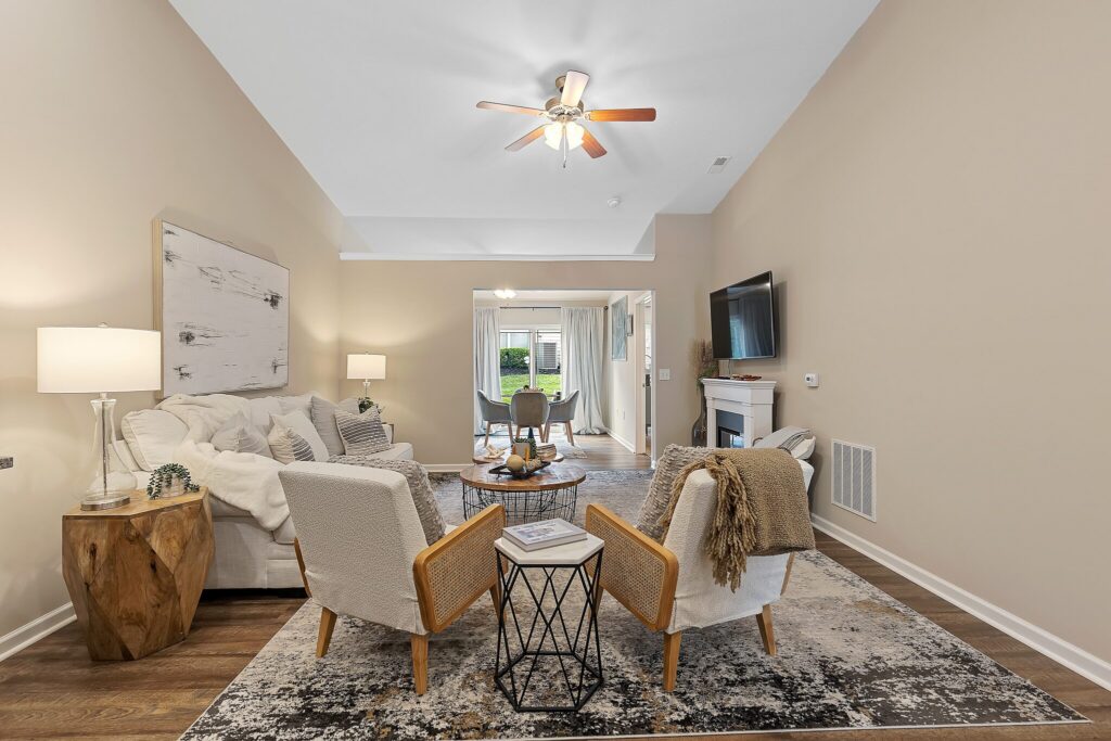 A modern living room with beige walls, white furniture, a rug, wall art, a ceiling fan, and a TV, opening to a dining area with large windows and patio access.