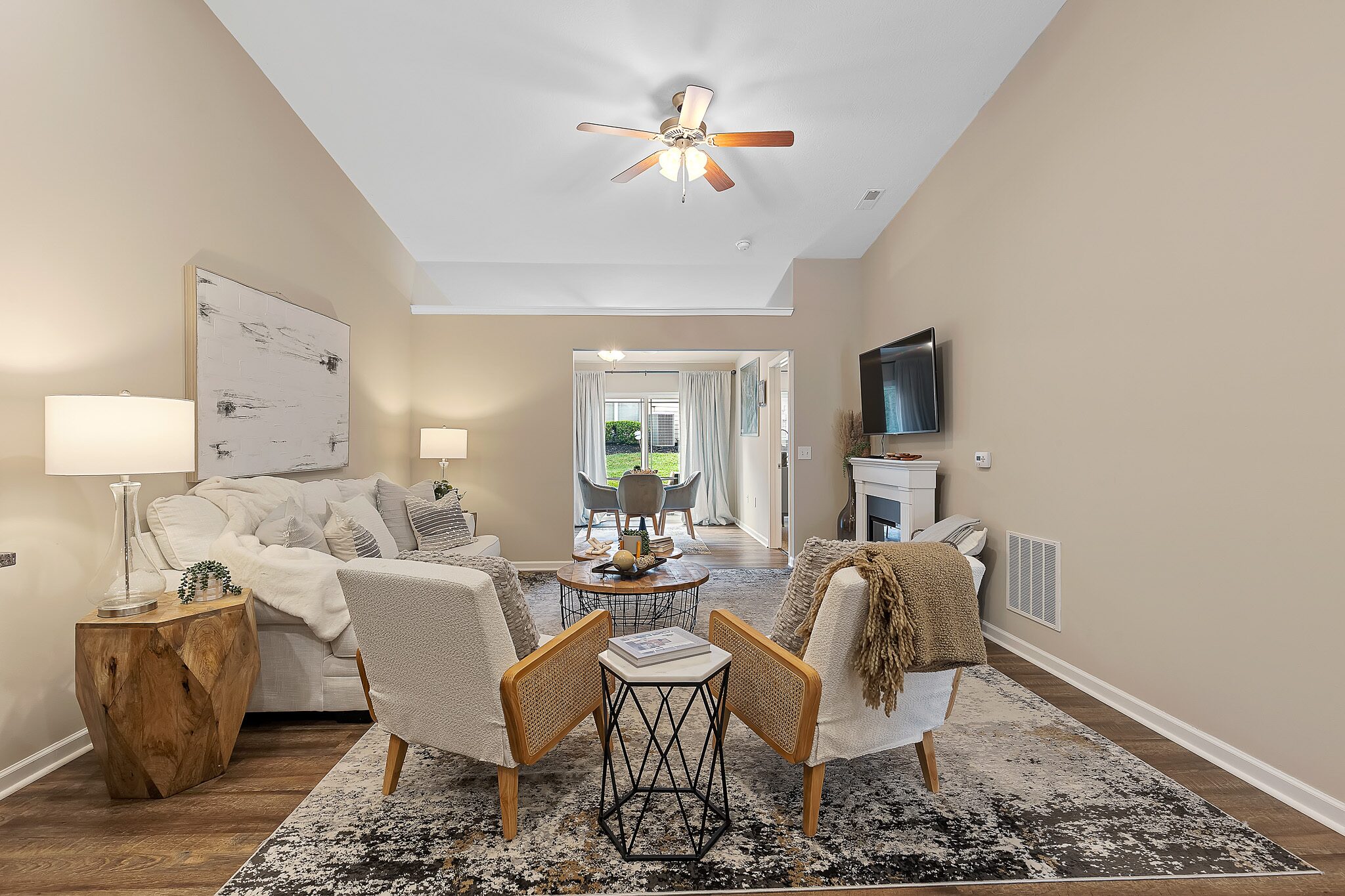 A modern living room with beige walls, white furniture, a rug, wall art, a ceiling fan, and a TV, opening to a dining area with large windows and patio access.
