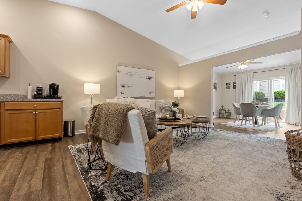 A cozy living room with neutral decor, armchairs, a round coffee table, and an adjacent dining area with natural light from large windows.