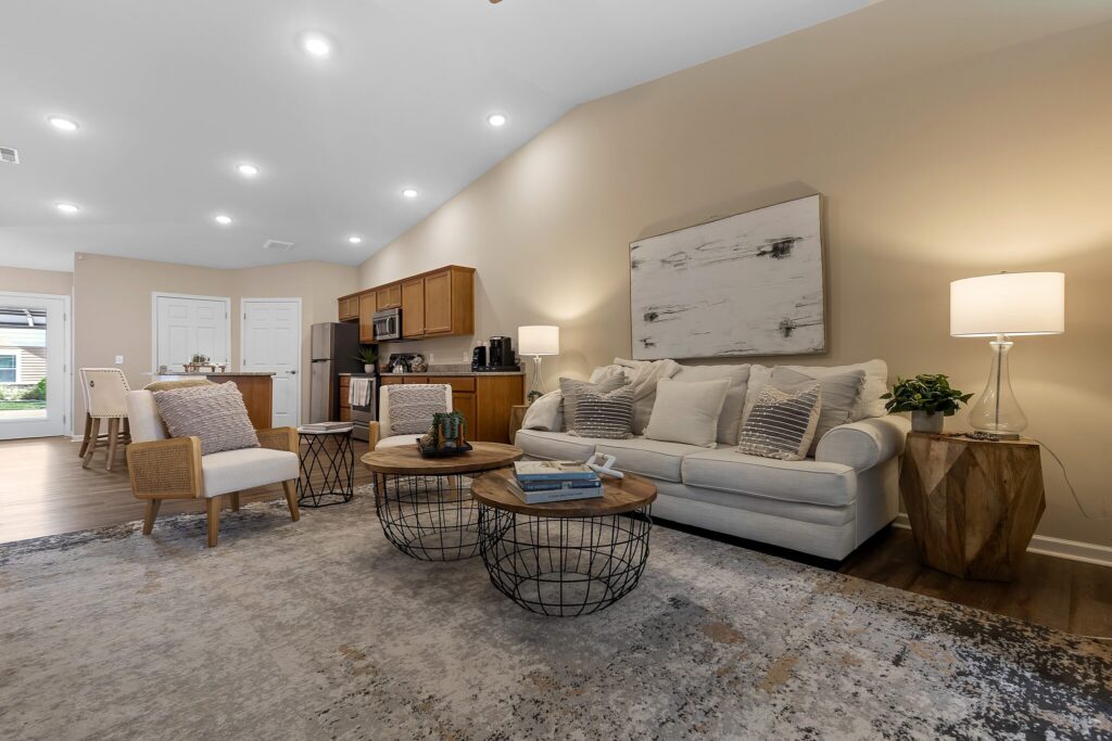 Modern living room with a beige sofa, accent chairs, round coffee tables, and neutral decor, open to a kitchen with wood cabinets and stainless steel appliances.