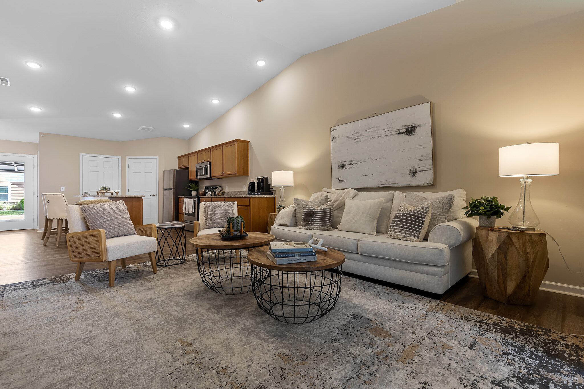 Modern living room with a beige sofa, accent chairs, round coffee tables, and neutral decor, open to a kitchen with wood cabinets and stainless steel appliances.
