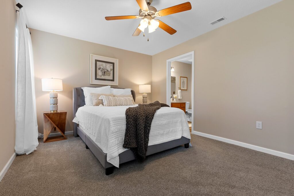 A bedroom with a neatly made bed, beige walls, carpeted floor, ceiling fan, two bedside lamps, and an open doorway leading to another room.