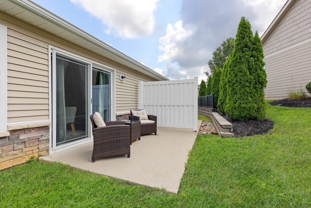 Small concrete patio with two brown wicker chairs and a side table, bordered by a sliding glass door, a privacy fence, green grass, and shrubbery.
